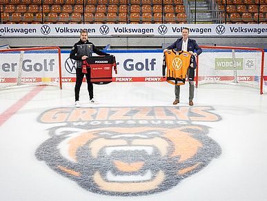 Zwei Männer stehen auf einem Eishockeyfeld und halten Sport-Trikots hoch.