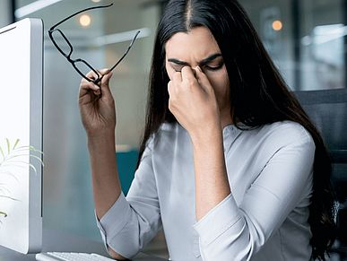 Frau sitzt vor ihrem Computer. Sie hat die Brille in der einen Hand, mit der anderen fasst sie sich an den Kopf. 