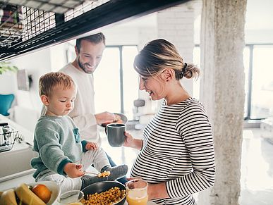Eine schwangere Mutter und ihr Mann stehen in der Küche. Ein kleiner Sohn sitzt auf dem Küchentresen und schaut sich Essen in der Pfanne an. Die Eltern lächeln.