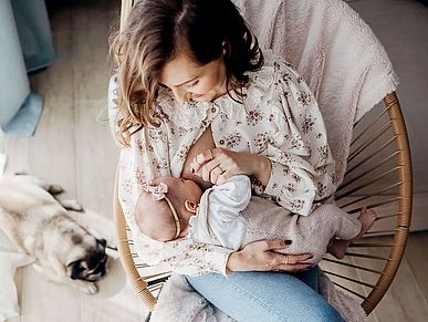 Eine Mutter stillt ihr Baby. Sie sitzt dabei in einem Korbstuhl, an dessen Fuß ein kleiner Mops liegt. Mutter und Kind sind in Pastell gekleidet, über der Stuhllehne hängt eine pastellfarbene Decke. Alles ist aus der Vogelperspektive zu sehen.