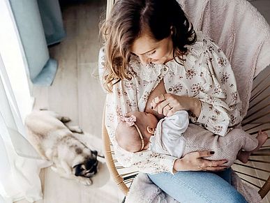 Eine Mutter stillt ihr Baby. Sie sitzt dabei in einem Korbstuhl, an dessen Fuß ein kleiner Mops liegt. Mutter und Kind sind in Pastell gekleidet, über der Stuhllehne hängt eine pastellfarbene Decke. Alles ist aus der Vogelperspektive zu sehen.