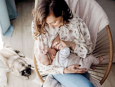 Eine Mutter stillt ihr Baby. Sie sitzt dabei in einem Korbstuhl, an dessen Fuß ein kleiner Mops liegt. Mutter und Kind sind in Pastell gekleidet, über der Stuhllehne hängt eine pastellfarbene Decke. Alles ist aus der Vogelperspektive zu sehen.