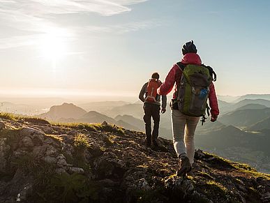 Zwei Personen mit Rucksäcken wandern auf einem bergigen Pfad. Der Boden ist mit Geröll bedeckt. Die Sonne scheint.