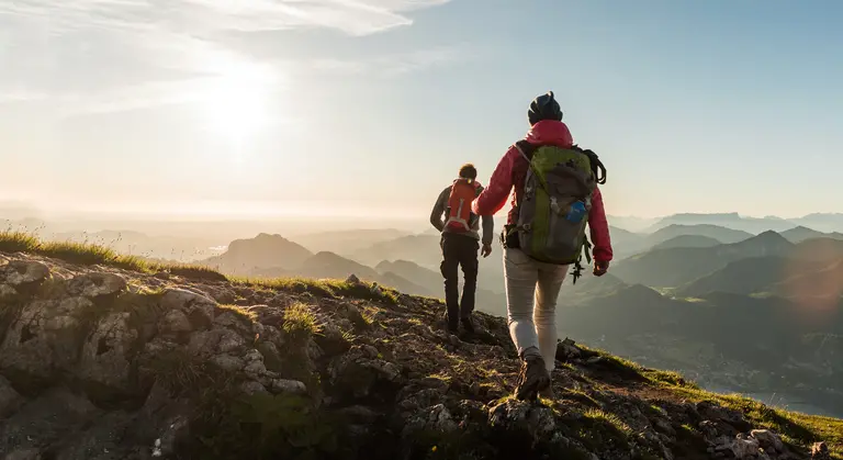 Zwei Personen mit Rucksäcken wandern auf einem bergigen Pfad. Der Boden ist mit Geröll bedeckt. Die Sonne scheint.