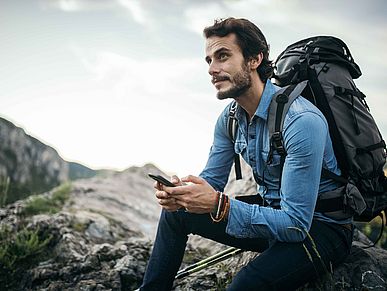 Mann mit Rucksack sitzt auf einem Berg und hält sein Smartphone in der Hand