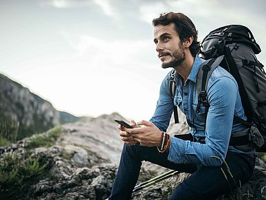 Mann mit Rucksack sitzt auf einem Berg und hält sein Smartphone in der Hand