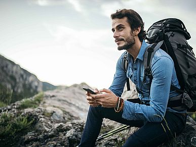 Mann mit Rucksack sitzt auf einem Berg und hält sein Smartphone in der Hand