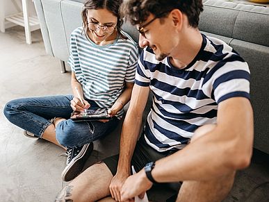 Zwei fröhliche junge Menschen sitzen auf dem Boden und bedienen ein Tablet.