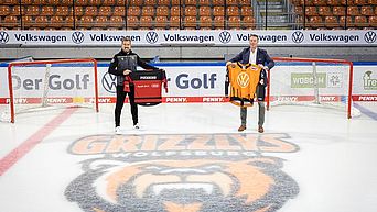 Zwei Männer stehen auf einem Eishockeyfeld und halten Sport-Trikots hoch.