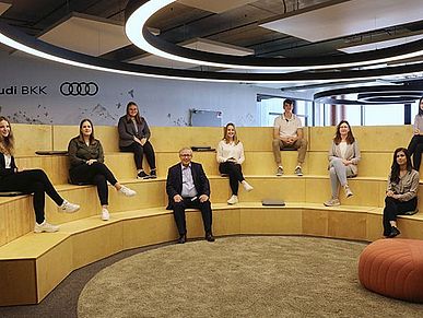 Eine Gruppe von Menschen sitzt in einem großen Raum auf einer Sitzgelegenheit.