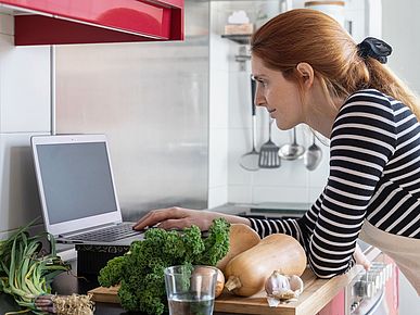 Frau steht in der Küche und schaut auf ihren Laptop. 