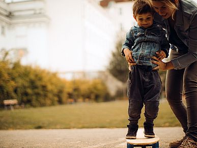 Ein kleiner Junge hält sich mit Hilfe seiner Mutter auf einem Skateboard