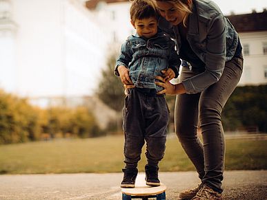 Ein kleiner Junge hält sich mit Hilfe seiner Mutter auf einem Skateboard